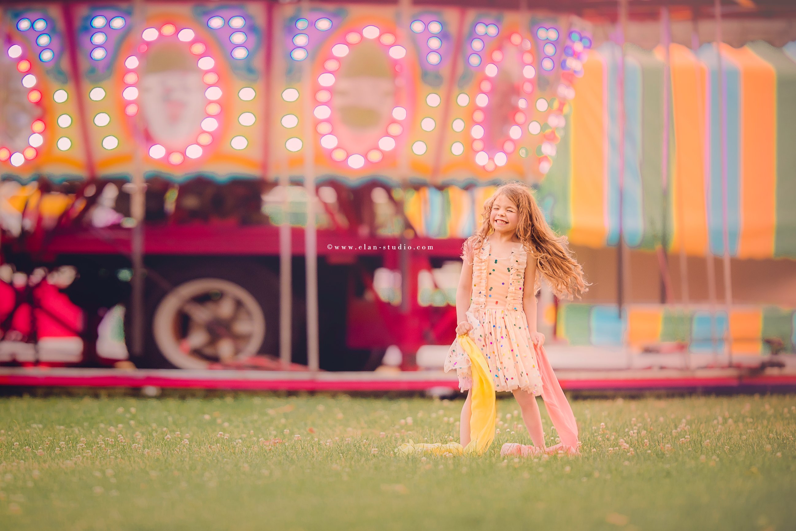 little girl with long hair, on grass at circus, with carousel behind her