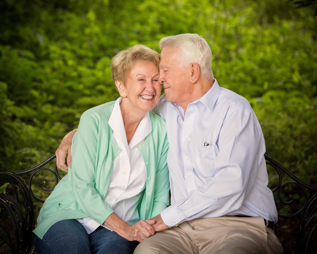 Elderly couple smiling and sitting closely on a bench in a lush green outdoor setting.