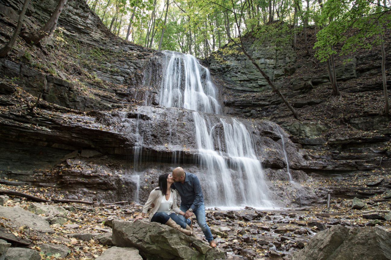 A couple sitting on rocks near a cascading waterfall in a forested area. Sherman Falls, Anaster