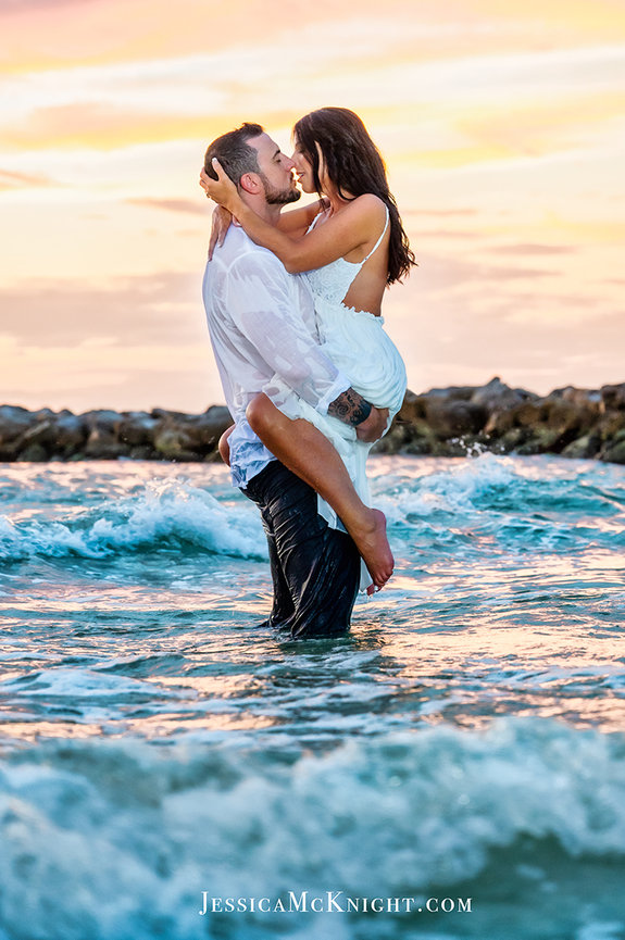 Couple kissing in the ocean at sunset, with waves around them and the sky lit in warm colors.