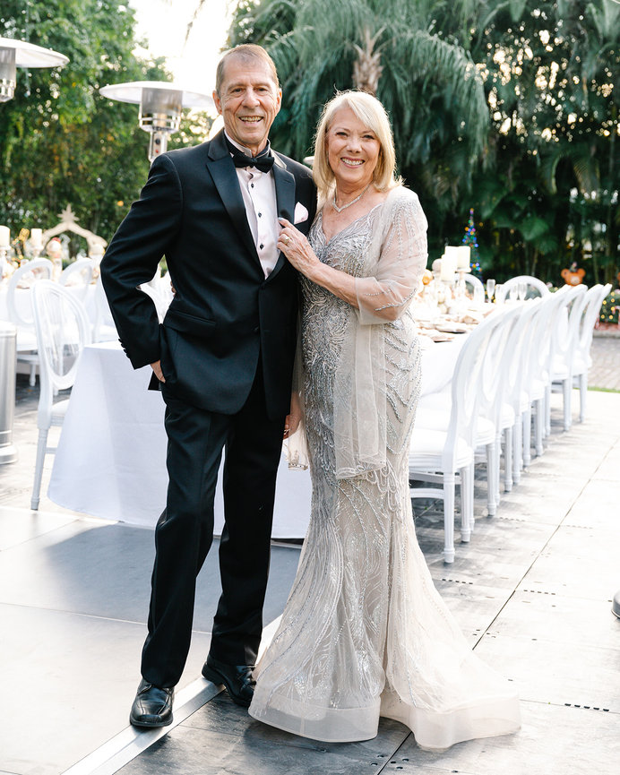Elegant couple in formal attire, smiling together at an outdoor event with tables set in the background.