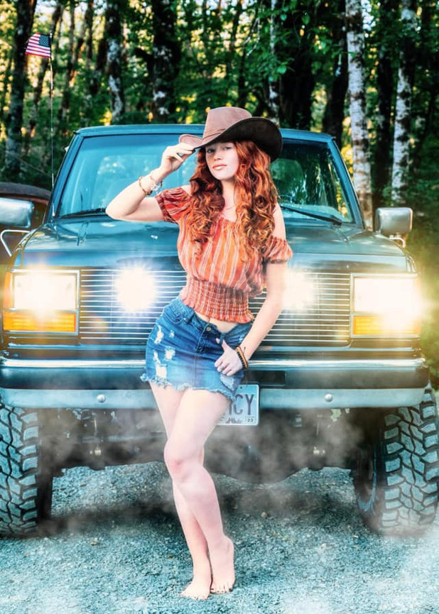 Woman in a cowboy hat poses by a 4x4 vehicle with bright headlights, set in a wooded area.