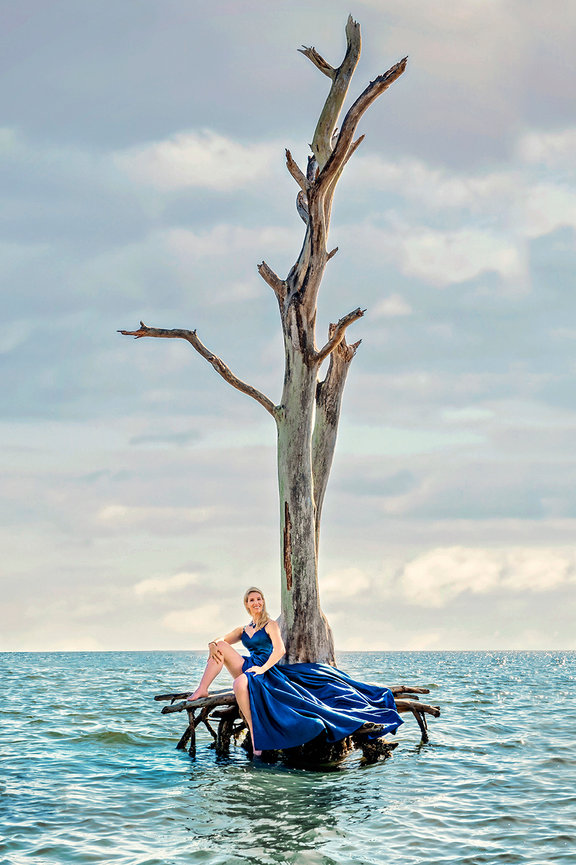 Woman in a blue dress sitting on a wooden structure in the water, beside a bare tree against a cloudy sky.