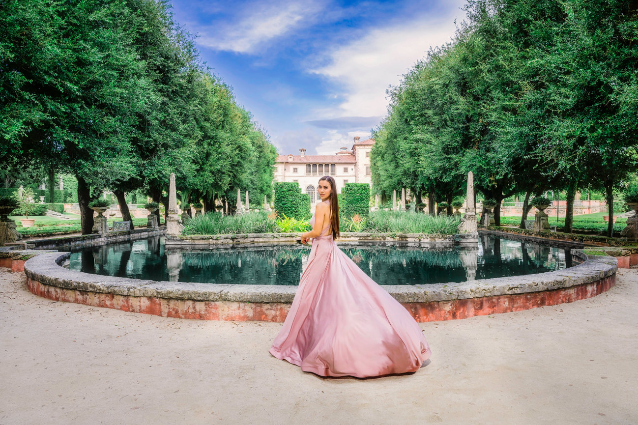 A woman in a flowing pink gown stands by a tranquil pond surrounded by lush greenery and a historic building in the background.