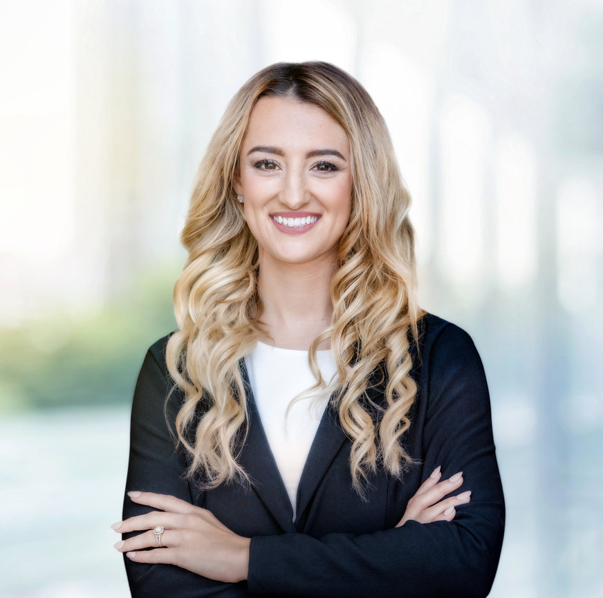 Smiling woman with long, wavy blonde hair in a black blazer, standing with arms crossed against a blurred background.