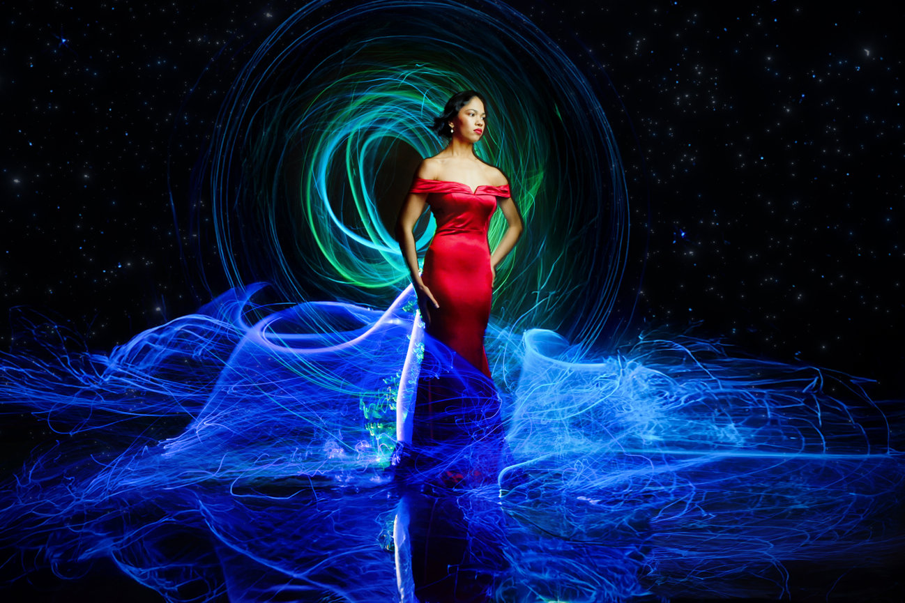 Woman in a red gown stands elegantly amid vibrant blue and green light trails against a starry background.