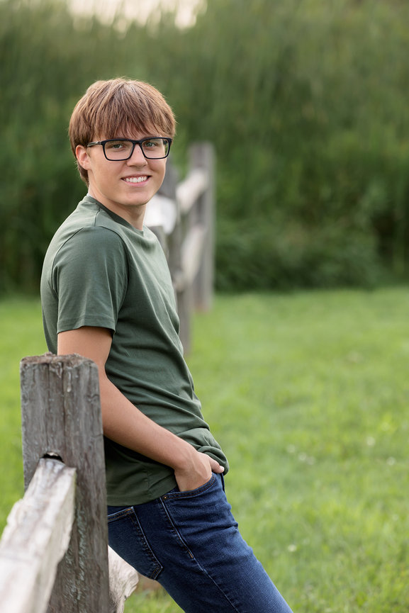 A person wearing glasses, green shirt, and jeans leans on a wooden fence with greenery in the background.