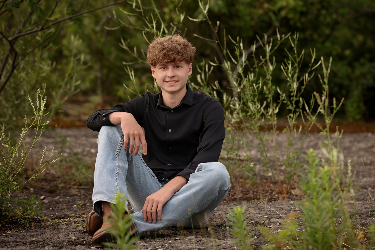 Young man sitting outdoors on rocky ground surrounded by green vegetation, wearing a black shirt and jeans.