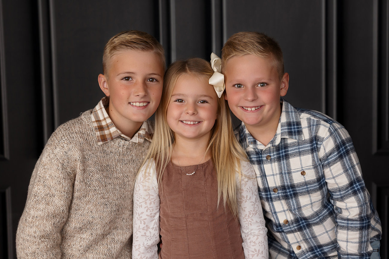 Three smiling children in casual outfits stand together in front of a dark paneled background.