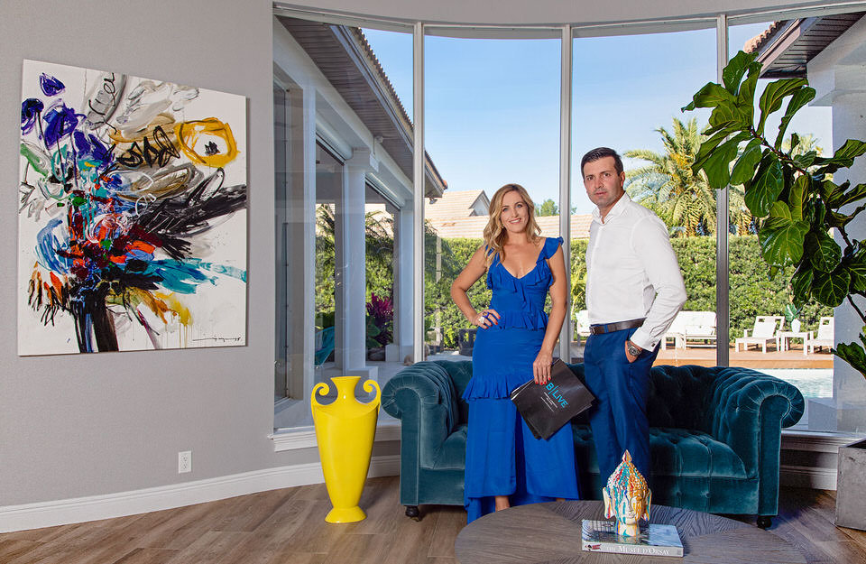 A couple in elegant attire stands in a modern living room with a large window, abstract art, and tropical plants.