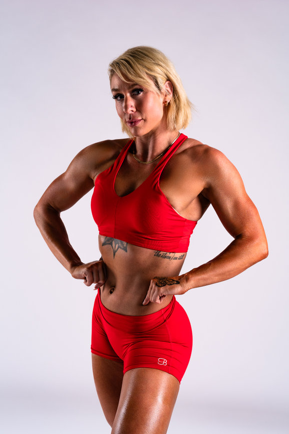 A muscular woman poses confidently in a red sports outfit in the studio of Austin Cinemaker Space.