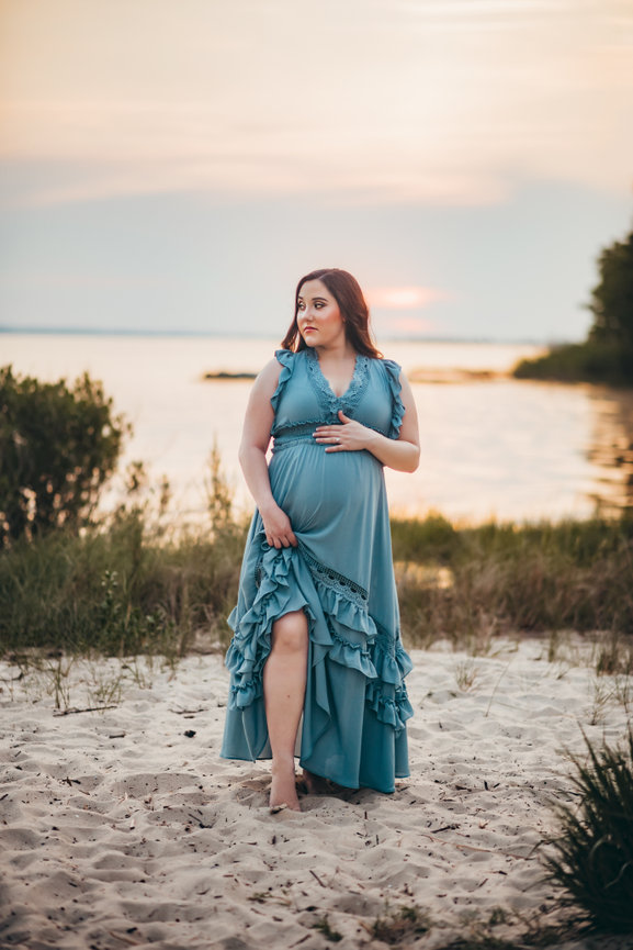 Pregnant woman in a blue dress standing on a sandy beach at sunset