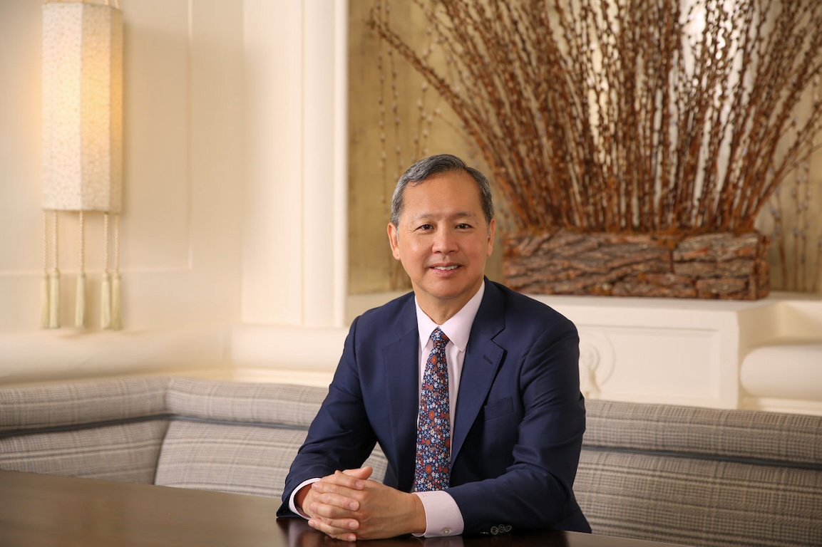 A person in a suit sits at a table, smiling, with a decorative background featuring tall, dried branches.