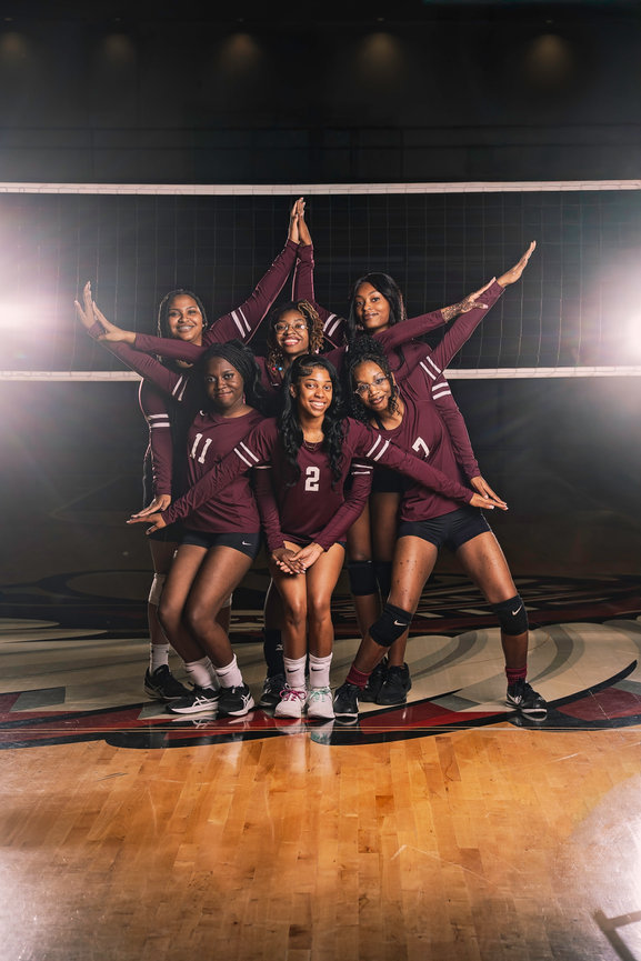Six female volleyball players in maroon uniforms pose together in a gym, showcasing teamwork and camaraderie.