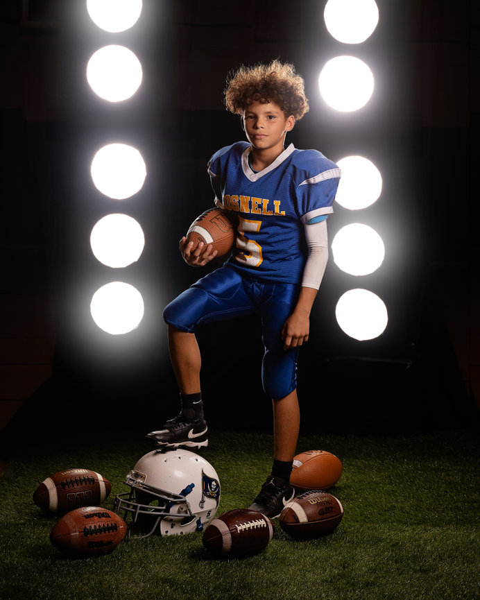 Young football player in blue uniform poses with footballs and helmet under bright lights.