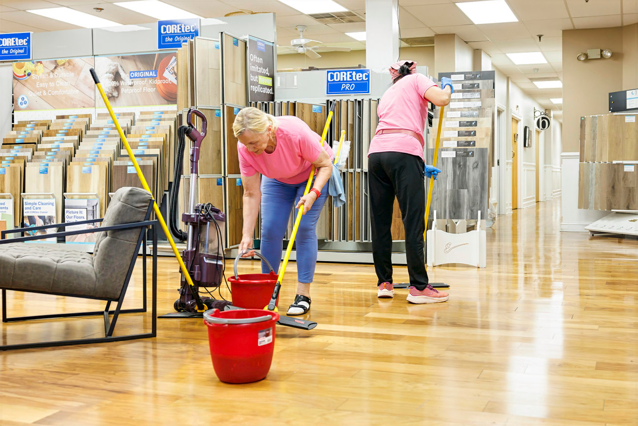 Two people cleaning a hardwood floor in a flooring store, using mops and red buckets.