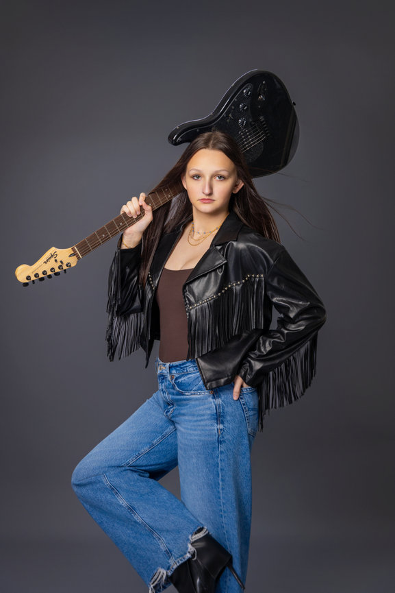 Studio portrait high school senior girl in jeans and black leather jacket with a guitar on her shoulder.