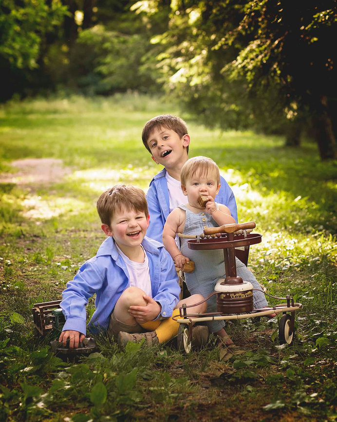 Three laughing brothers sit in the grass. The toddler brother is in an antique stroller.
