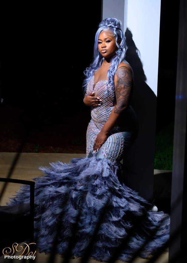 A woman in a detailed, feathered gown, with long hair, stands gracefully under a spotlight.