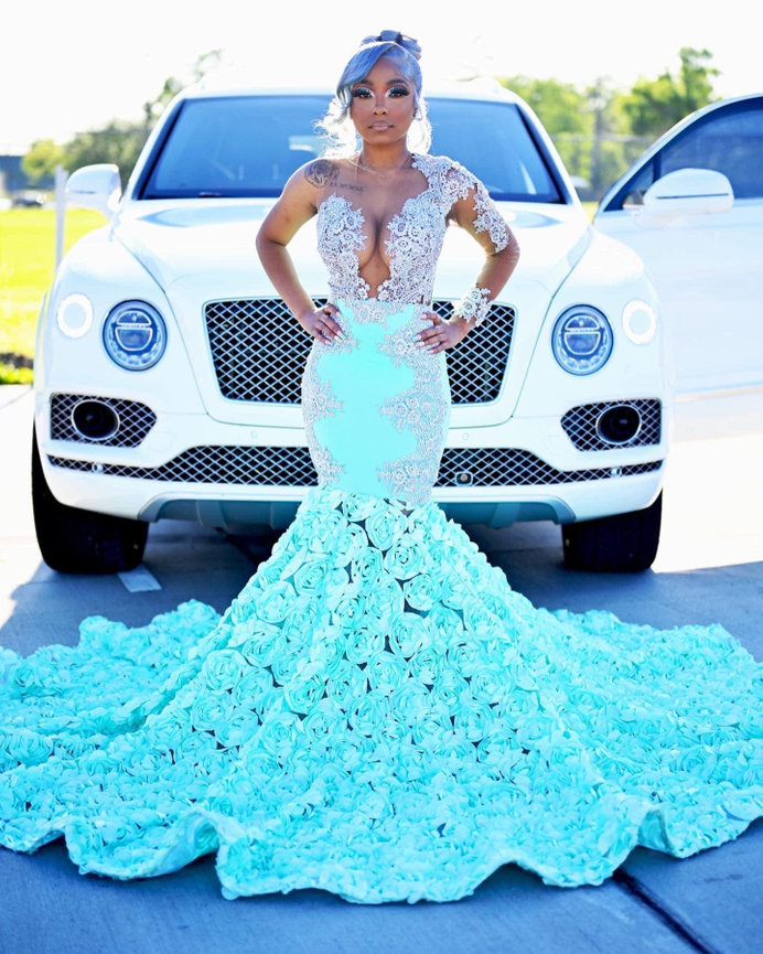 Woman in ornate turquoise floral gown stands in front of a white luxury car, outdoors.