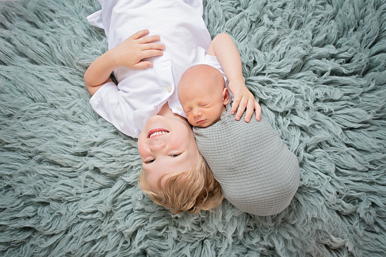 Sibling joy: Young child lying on a fluffy rug, smiling, with a newborn baby wrapped in a blanket.