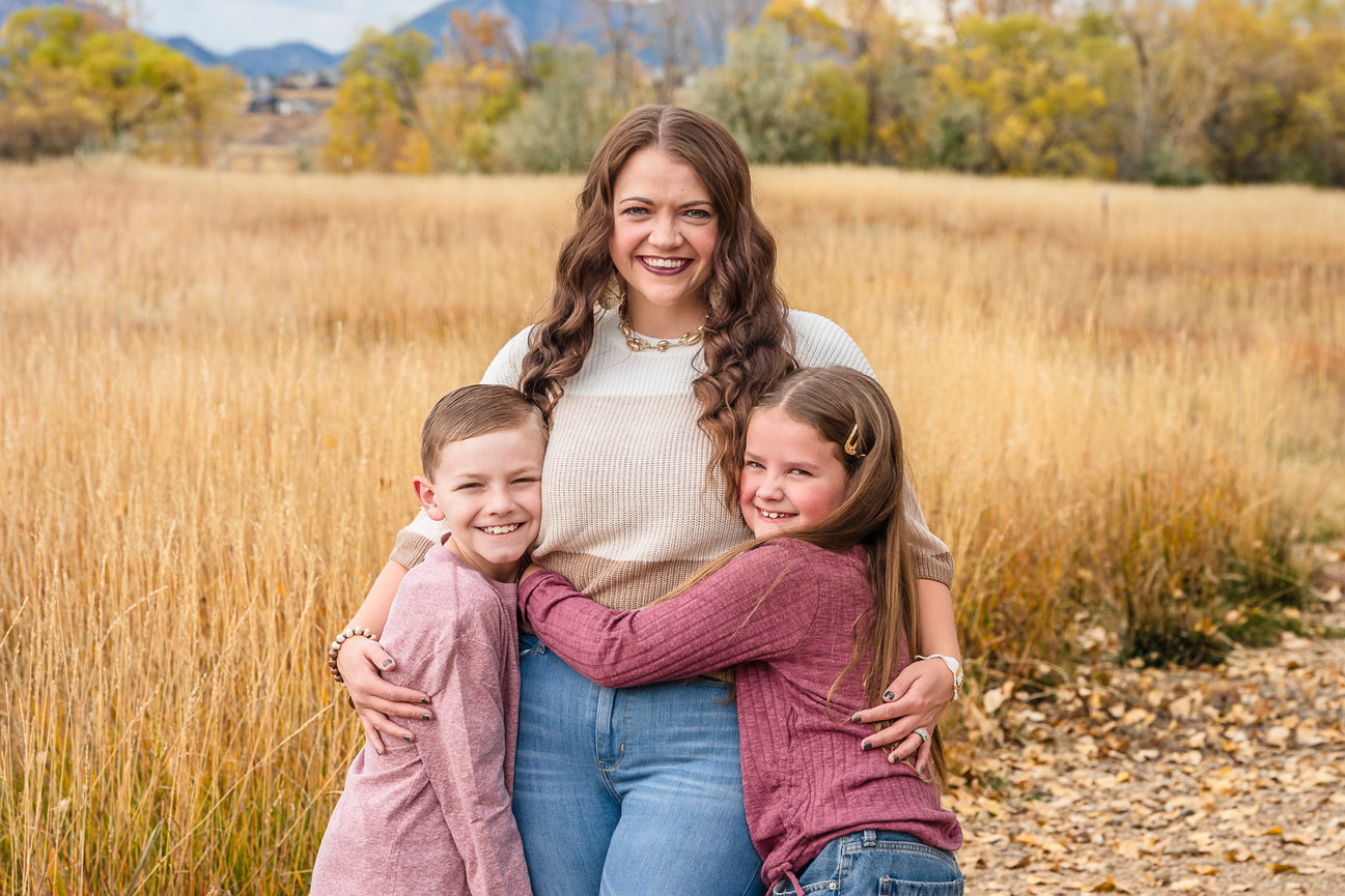A mom stands in a grass field. Her children are snuggled close with big smiles.