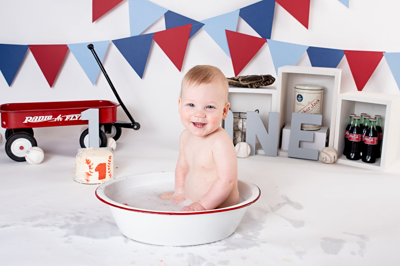 Baby smiling while bathing in a white tub, surrounded by red and blue decorations, with a small red wagon nearby.