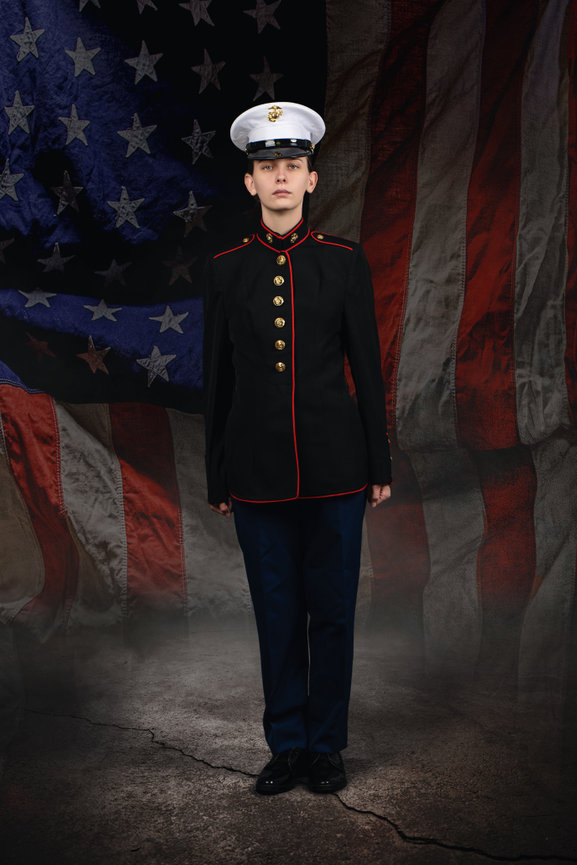 Person in military dress uniform standing before a faded American flag backdrop