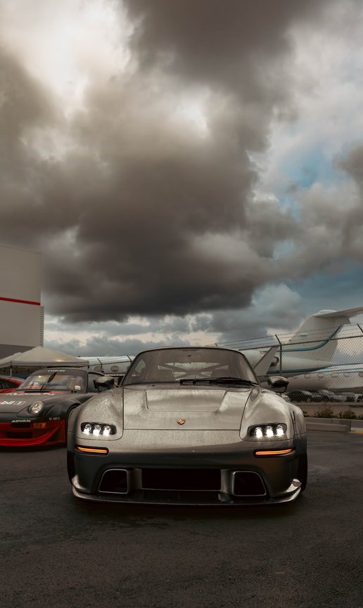 Low-angle view of a silver Porsche race car parked under dramatic storm clouds at an airfield