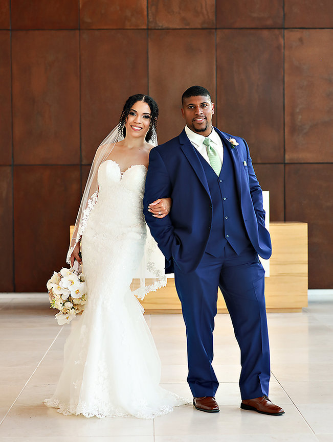 Bride in a lace gown with a bouquet and groom in a blue suit, smiling and standing together indoors.