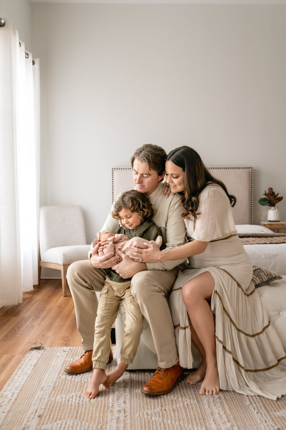 Family of four cuddling a newborn on a bed in a bright, minimalist bedroom