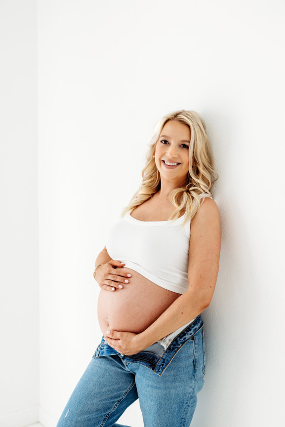 Pregnant woman smiling, wearing a white tank top and jeans, leaning against a white wall.