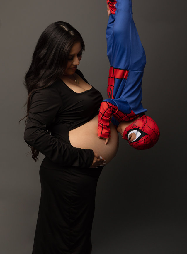 Pregnant woman in black dress with child in Spider-Man costume hanging upside down touching her belly.