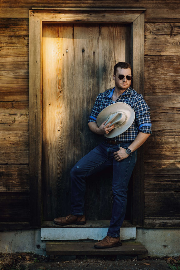Man in sunglasses leaning against a rustic wooden doorway holding a cowboy hat