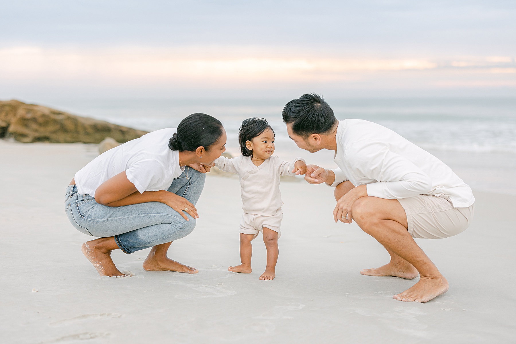 family kissing baby's hands on the ocean with pastel sky