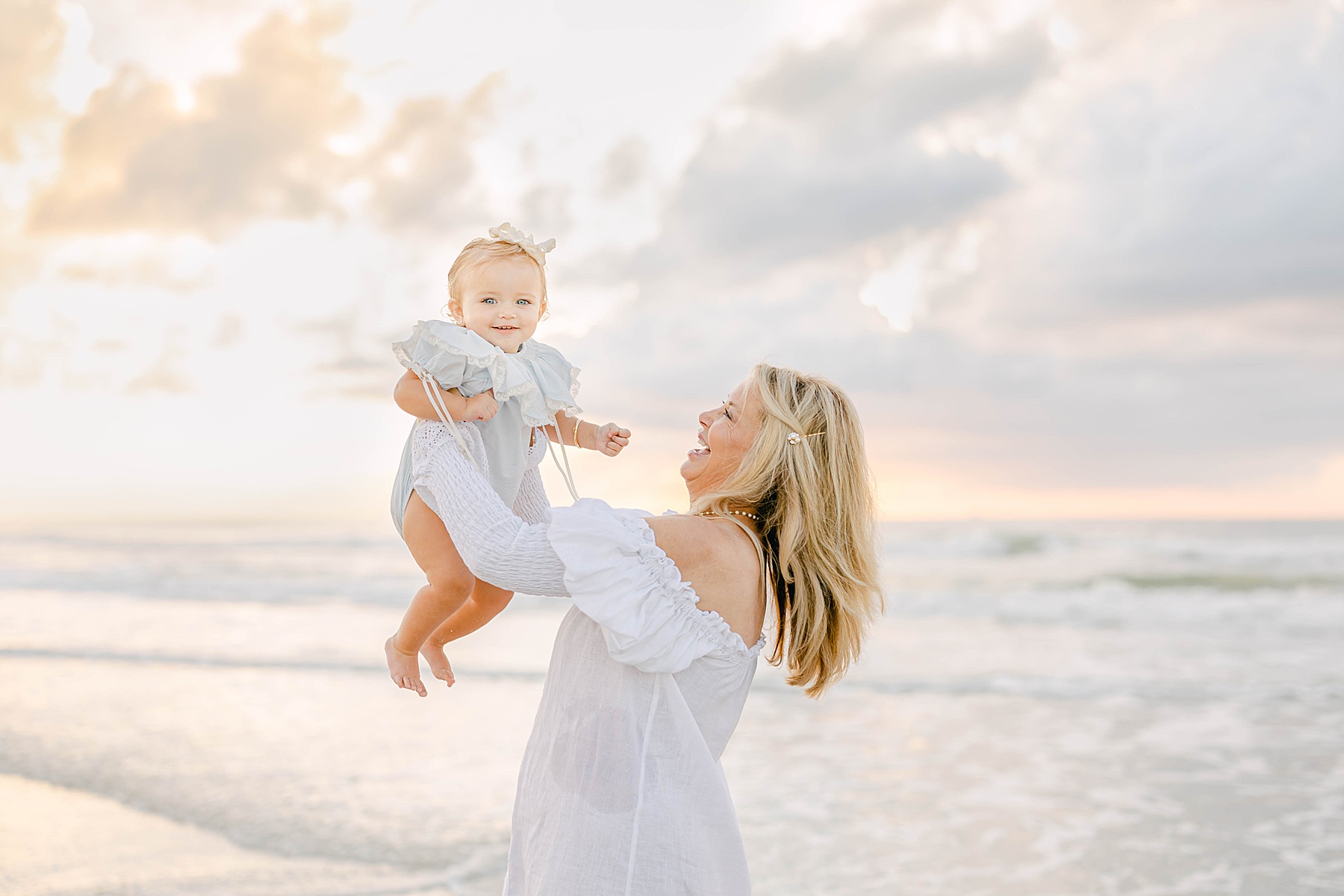 older woman in white dress holding baby in the water at the beach at sunrise