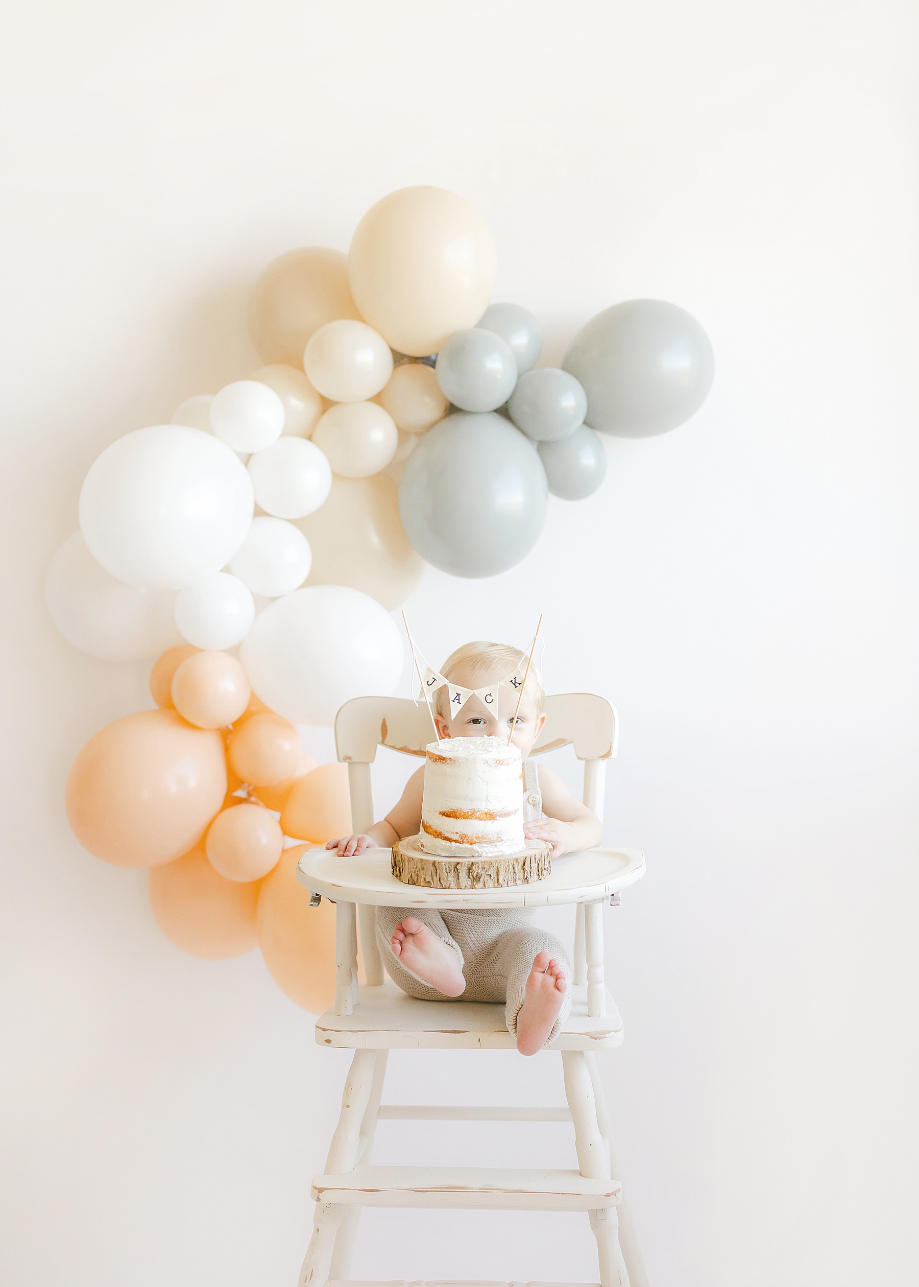 baby boy sitting in white high chair in front of white birthday cake and neutral balloons