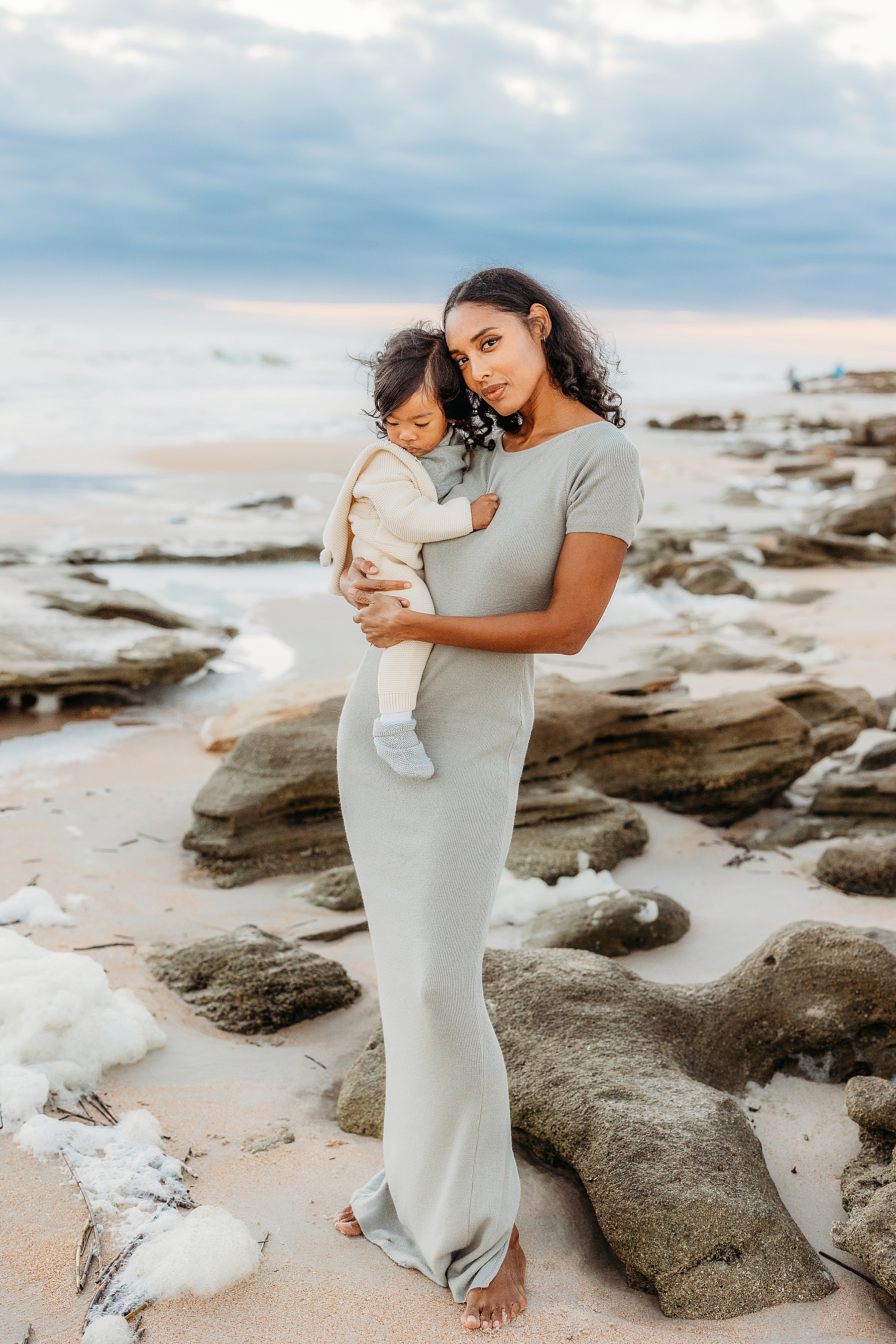 woman holding baby boy on the beach at sunrise wearing sage green long dress at sunrise