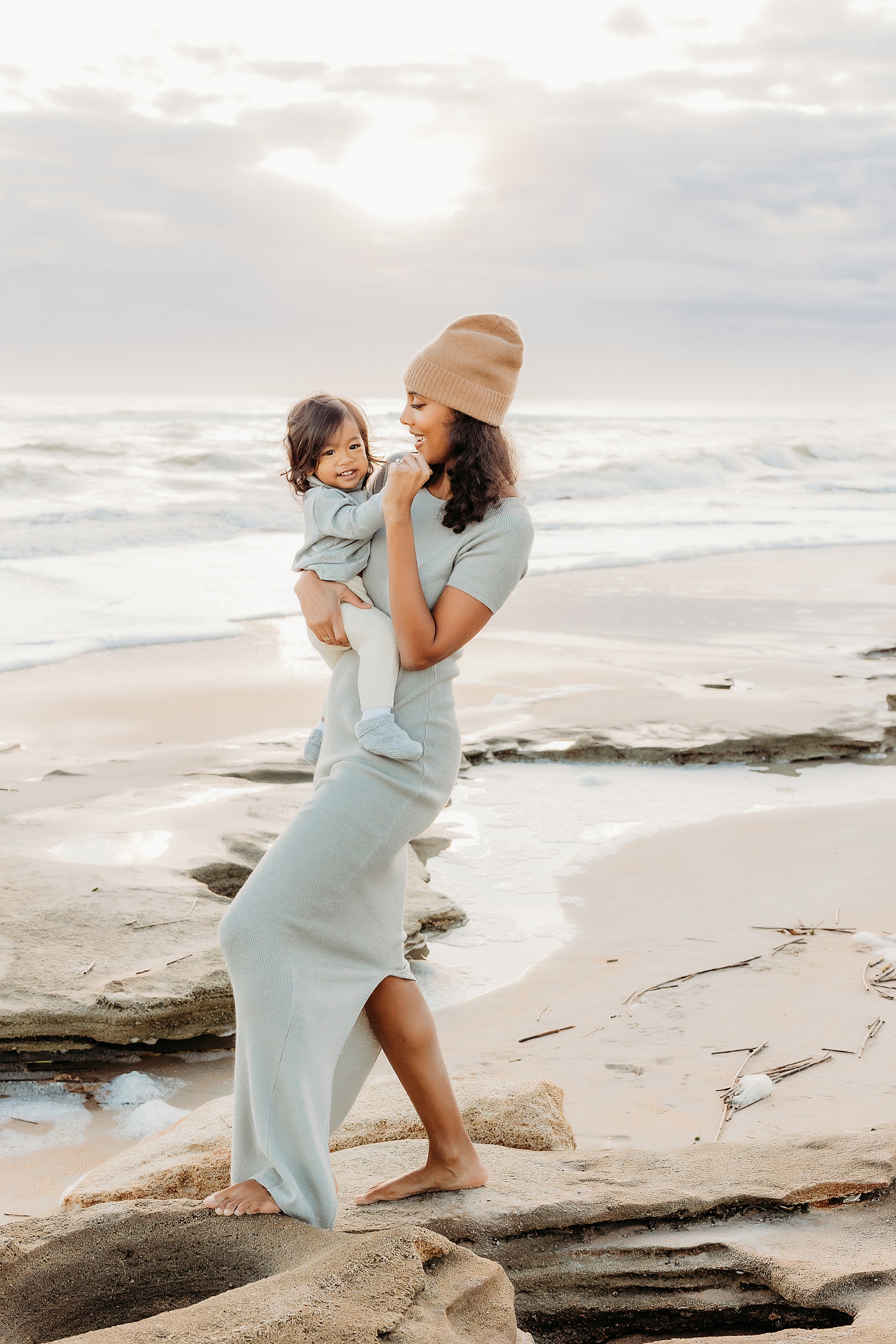 woman holding baby boy at sunrise on the beach wearing knit taupe hat