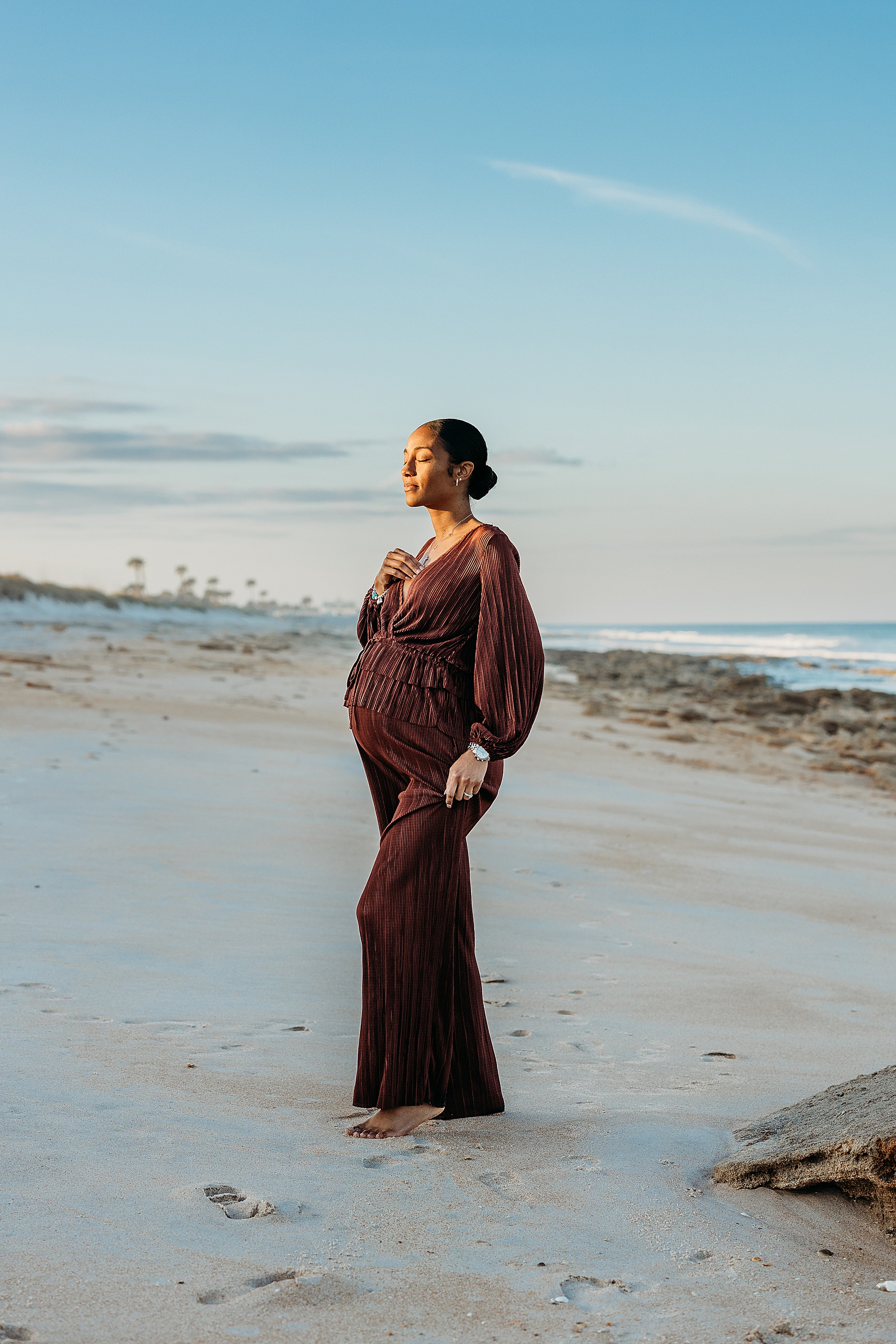 pregnant woman in dark jumpsuit standing on the beach in harsh sunlight