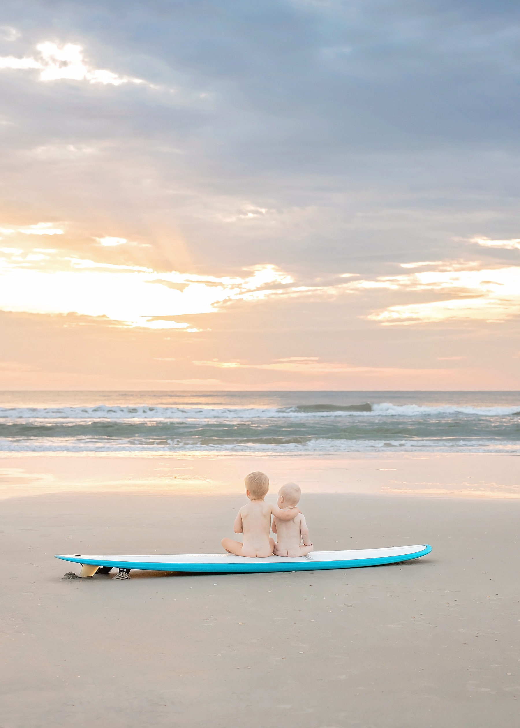 baby boys naked sitting on a surfboard at sunrise in st. augustine beach florida with pastel sky 