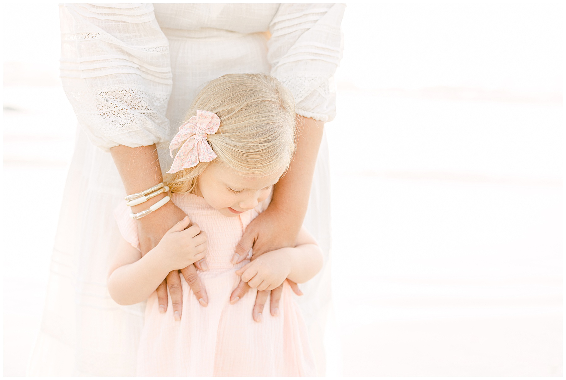 little blond haired girl dressed in pale pink dress holding onto mom on the beach at sunrise