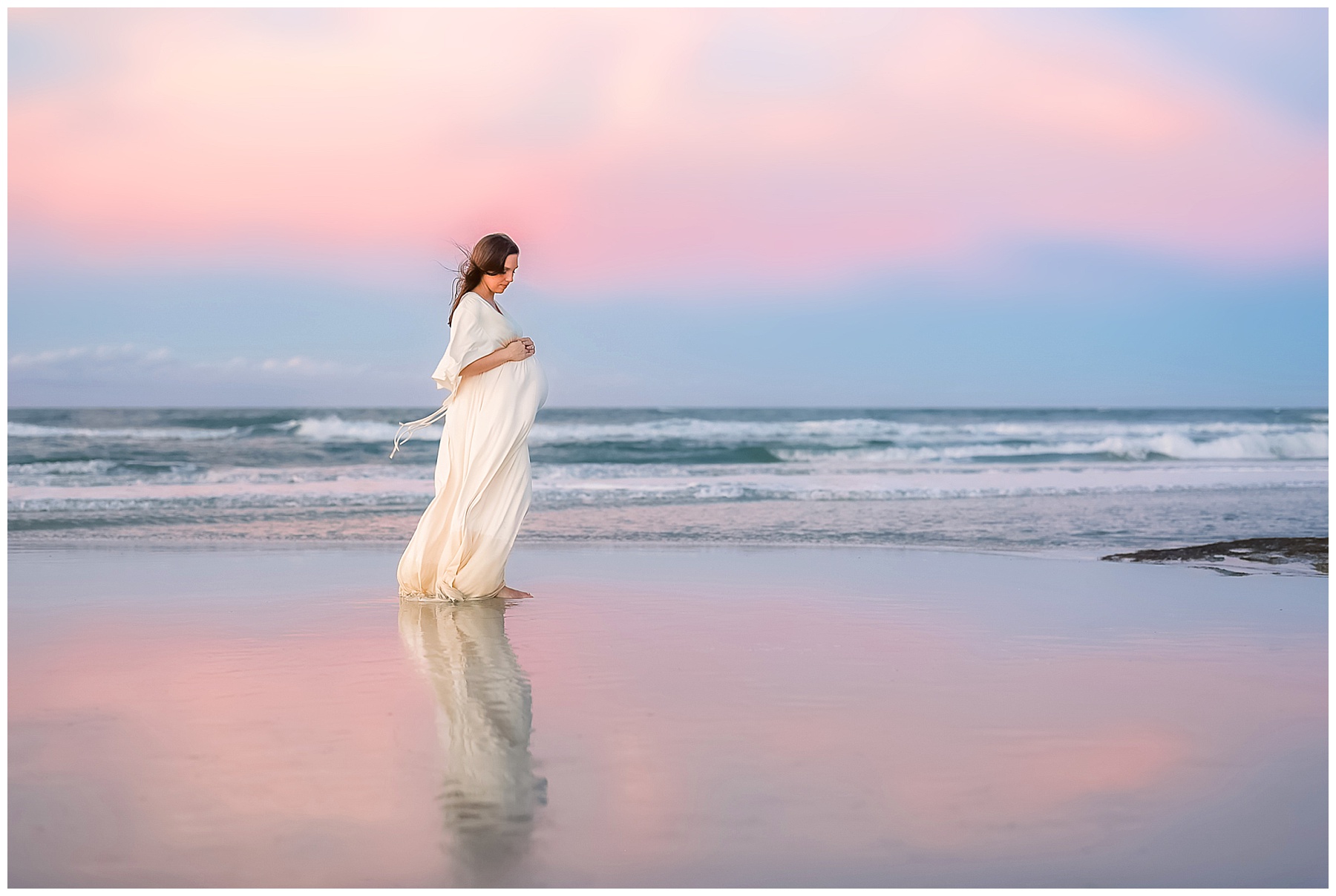 pregnant woman in long cream dress standing by the water at sunset with pastel skies