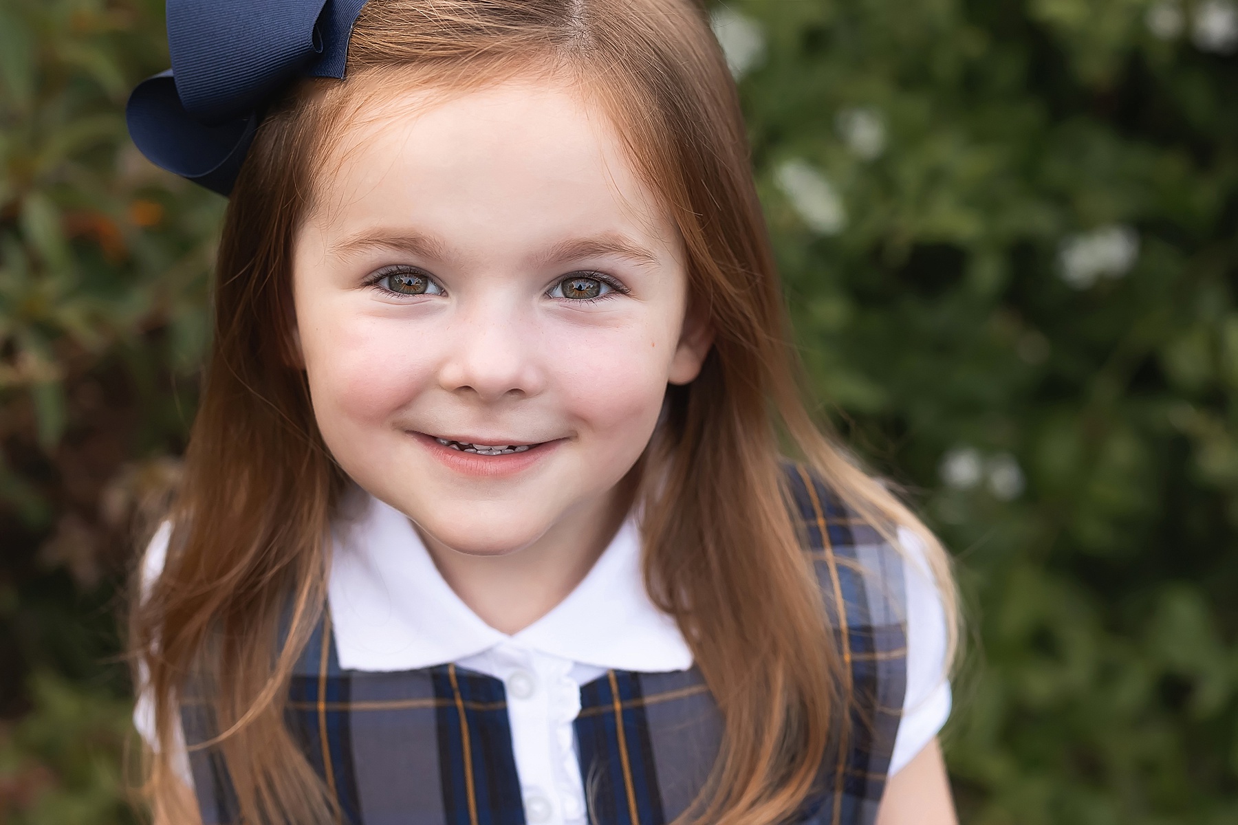 little girl with red hair dressed in navy and white plaid smiling in a garden on her first day of pre-school in st. augustine florida 
