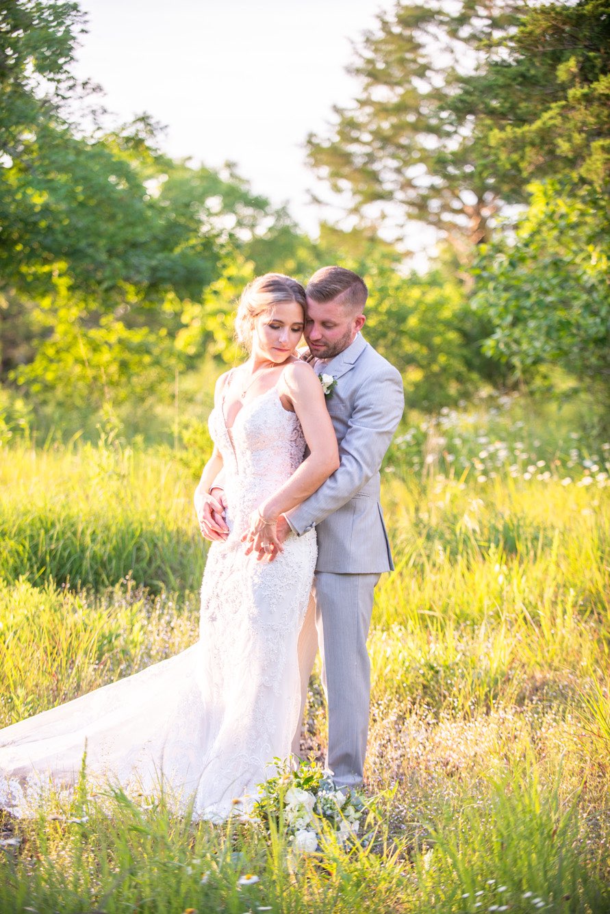 Bride wearing a lace boho gown and groom in a grey suit standing together in a field of flowers at sunset.