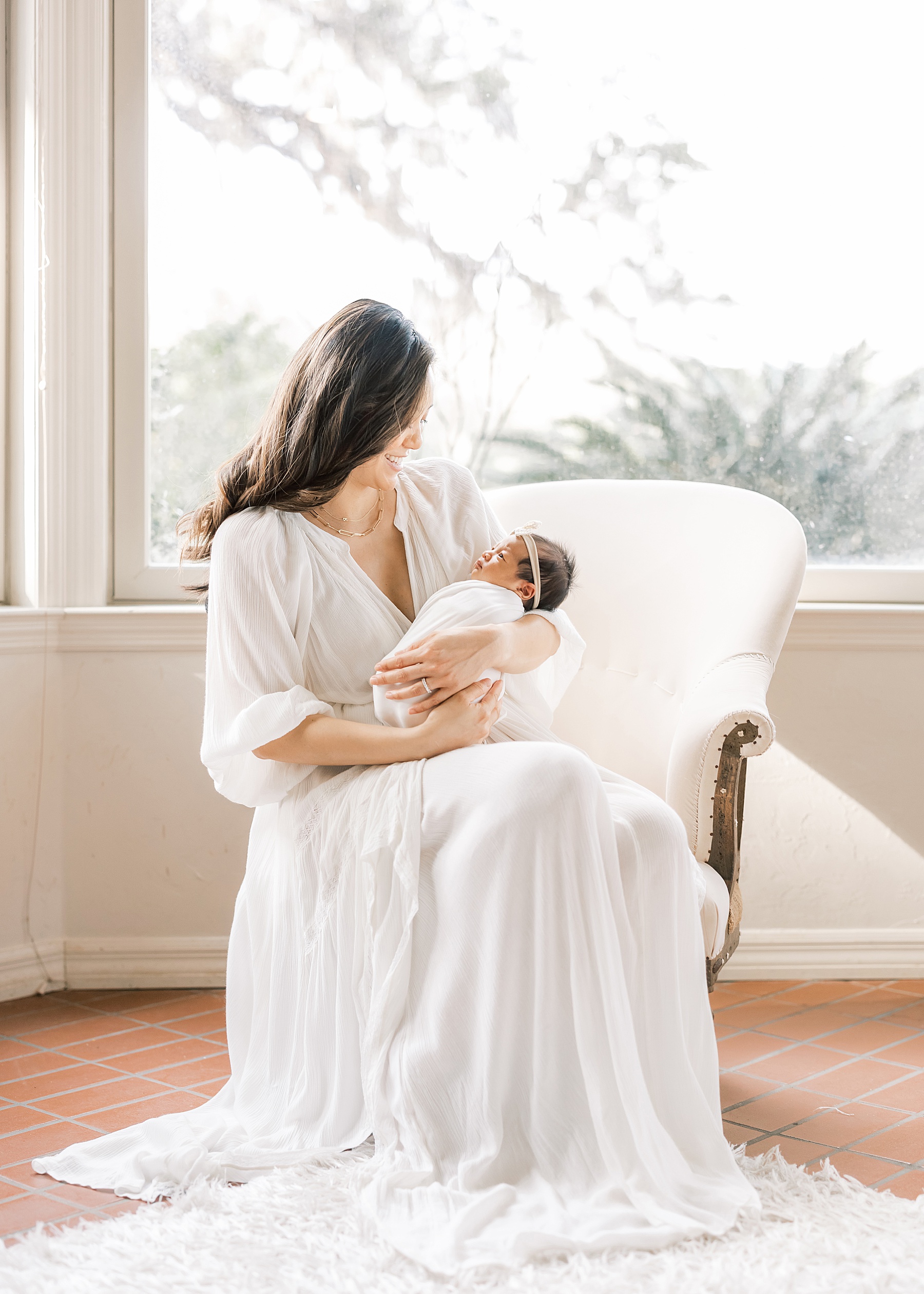 woman with long dark hair in long white dress sitting in vintage chair holding newborn baby girl