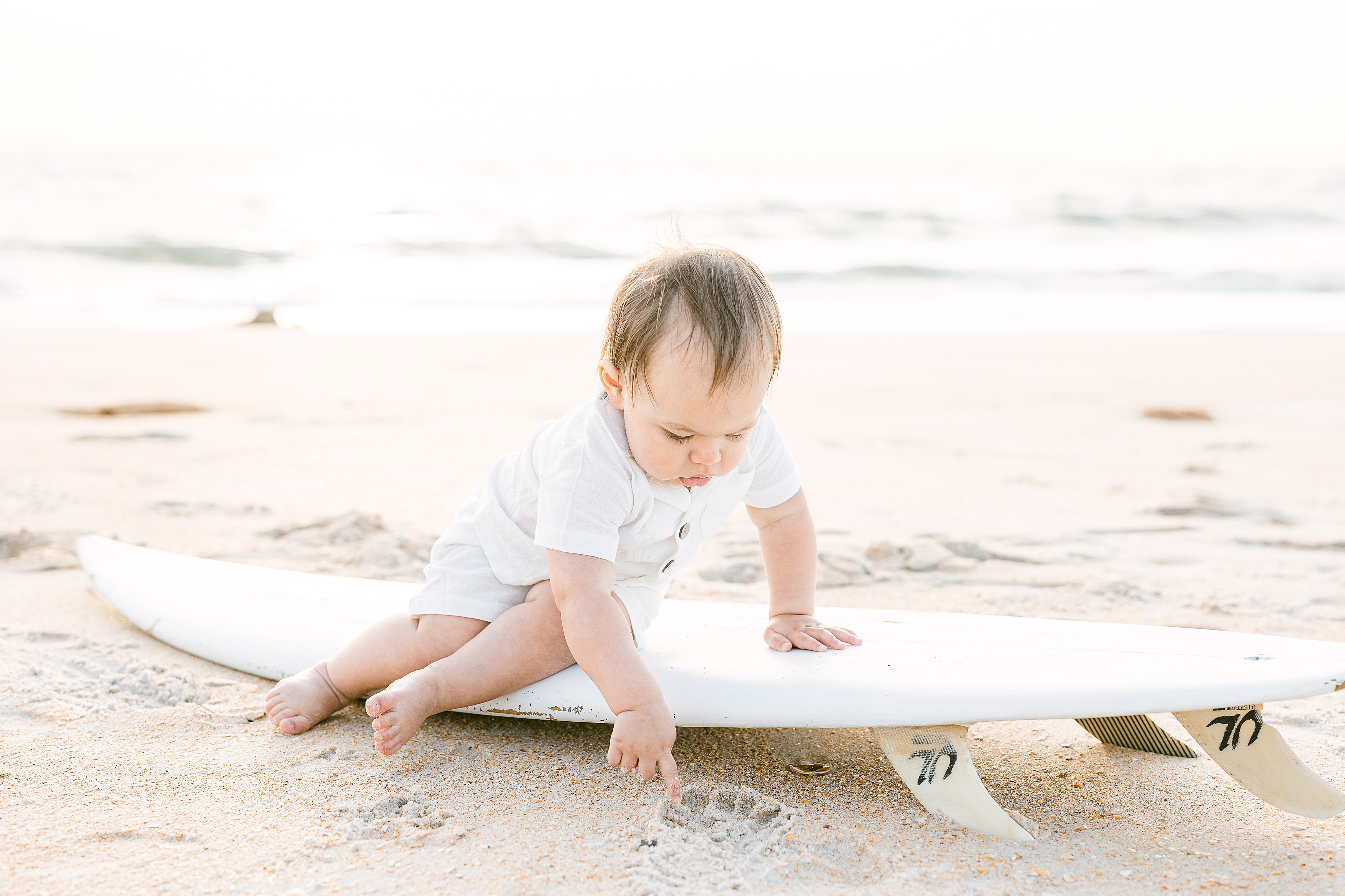 baby boy in linen overalls sitting on white surfboard on the beach at sunrise