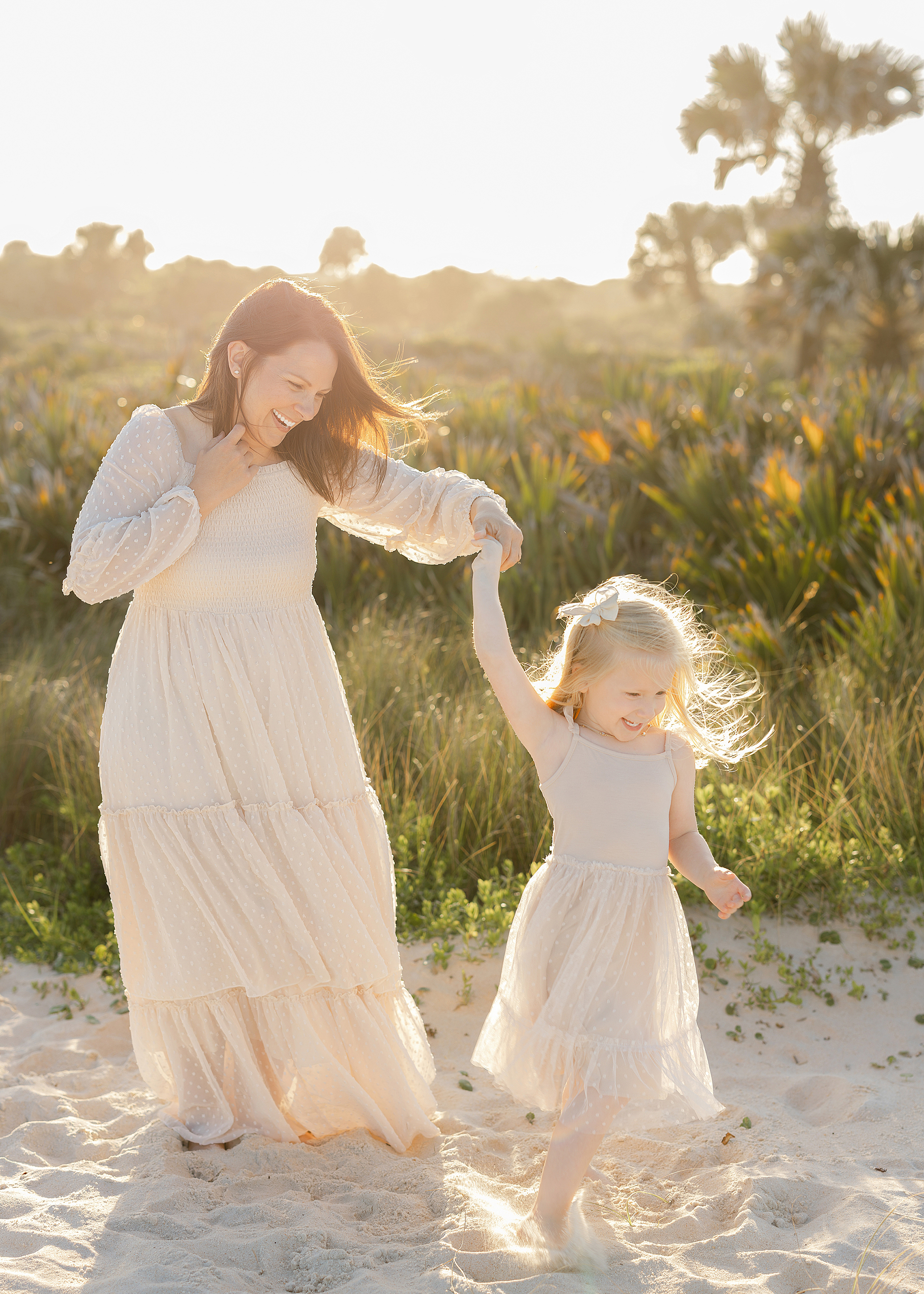 A little girl and her mother dressed in cream tulle dresses twirl together on the beach at sunset.