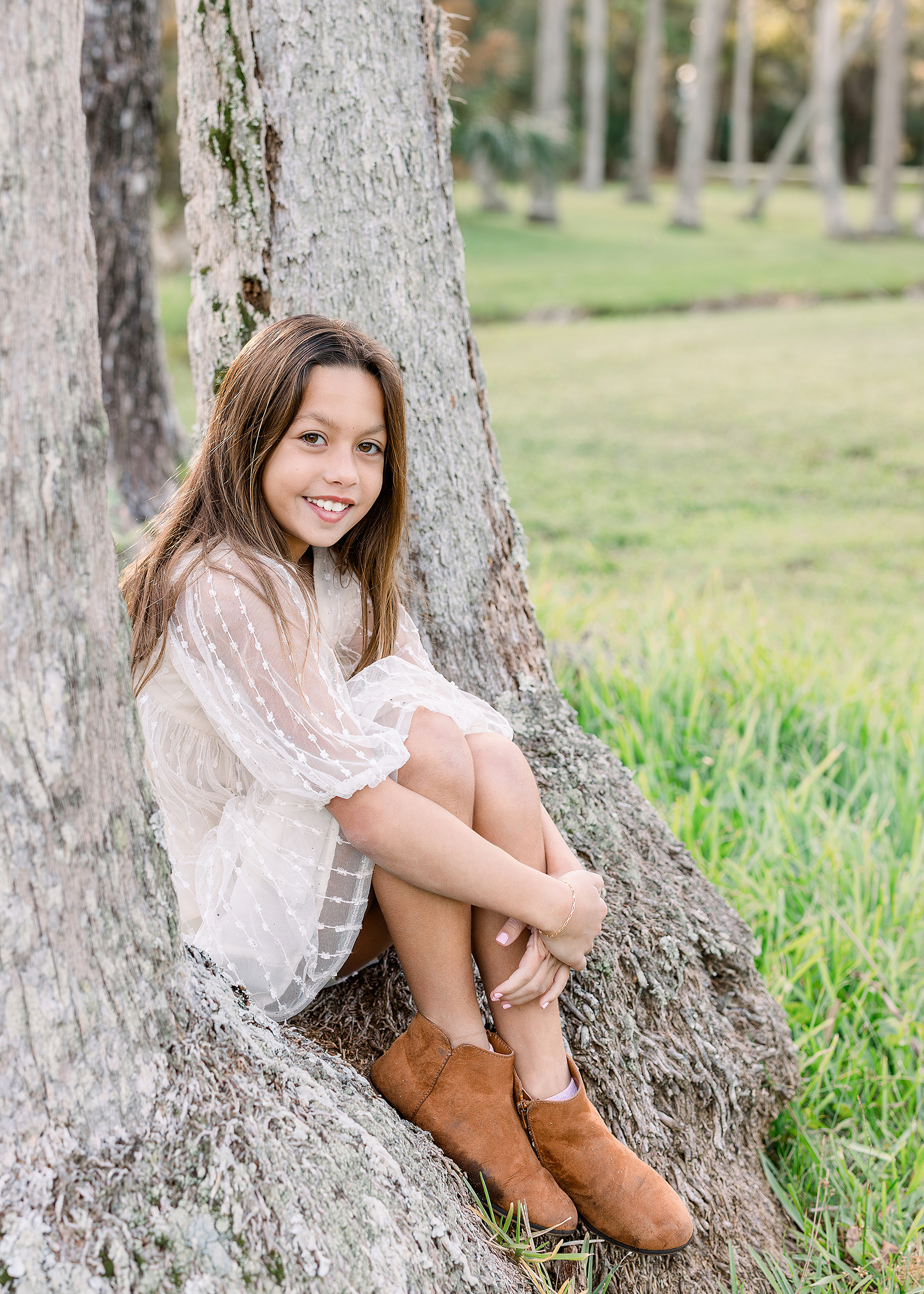 An airy portrait of a little girl in a cream dress with brown suede booties sits on a tree stump in Johansen Park in Atlantic Beach.