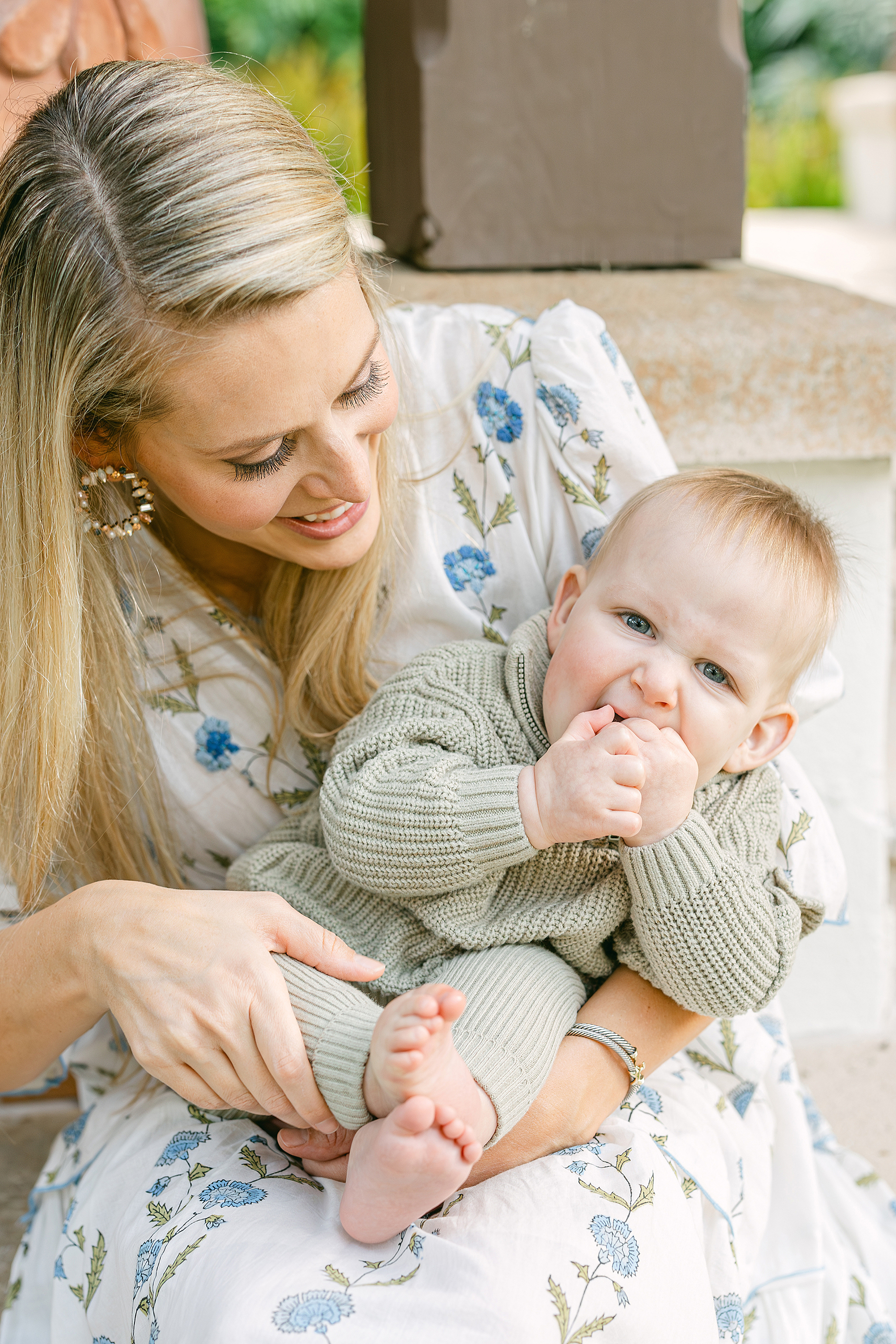 A baby boy with blue eyes in a sage green romper puts his foot in his mouth and sits with his mother.